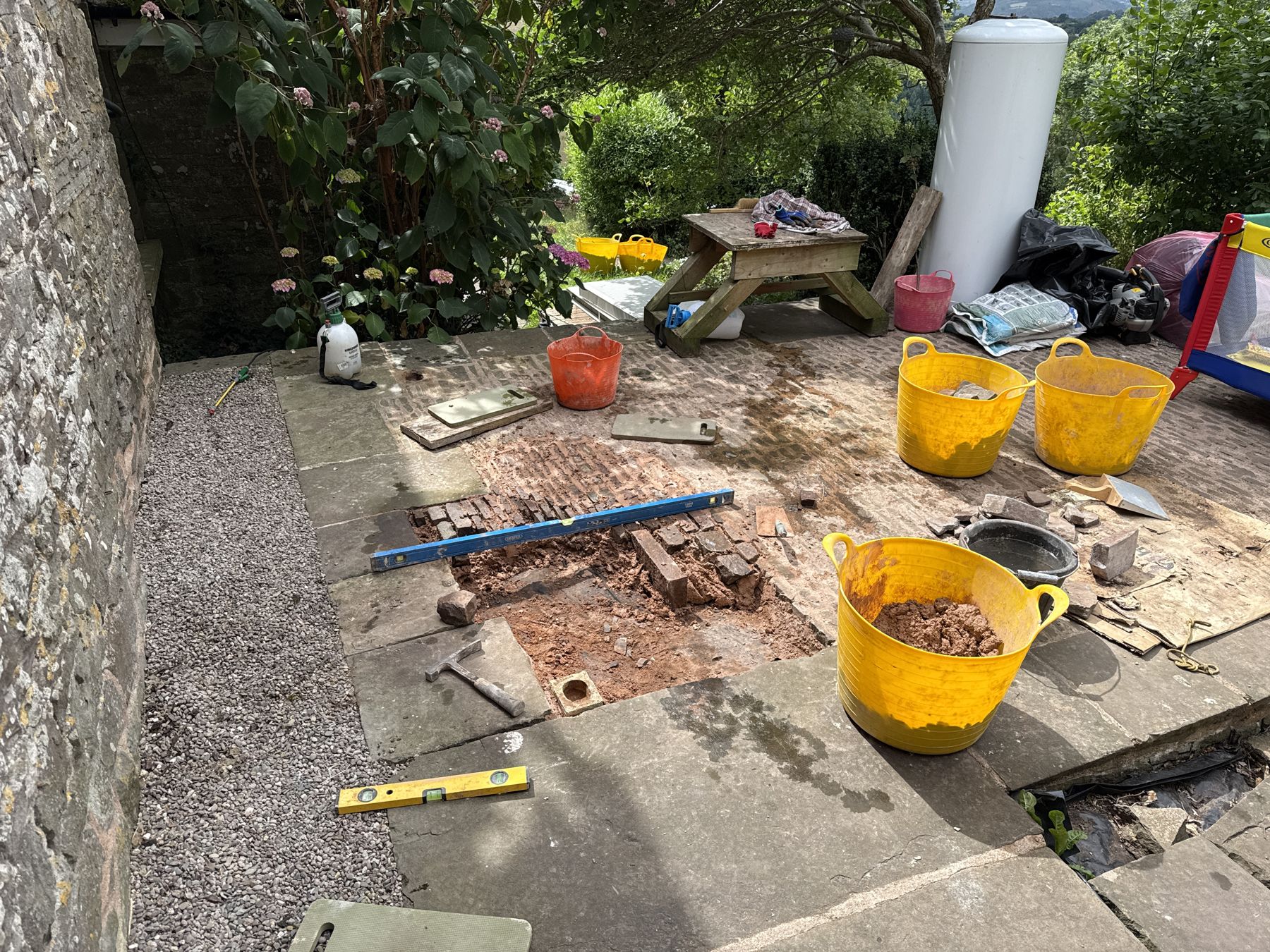 A section of the cobbled courtyard being relaid at Pengilfach in July 2025: the old cobbles lifted, sand and setts ready in yellow trug buckets, a spirit level laid across the bed