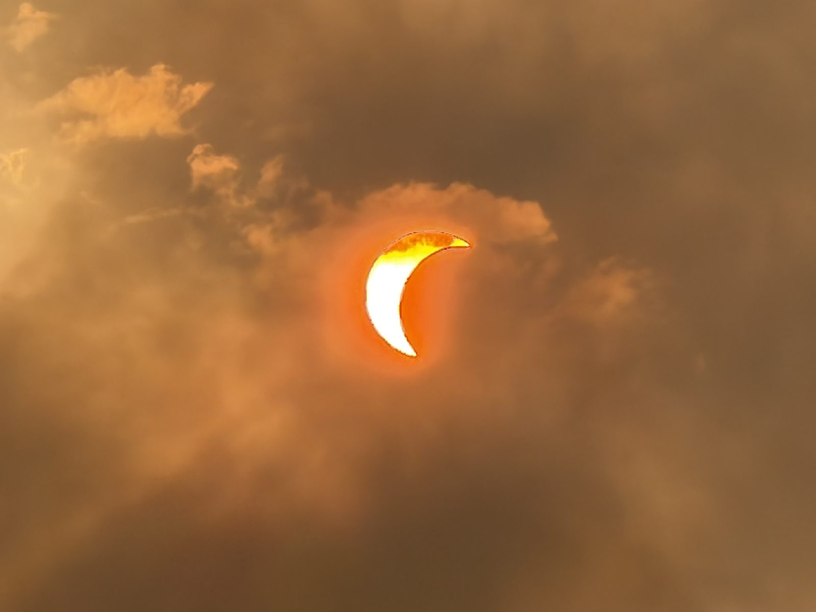 The eclipsed sun as a bright crescent with a glowing red-orange rim, framed by smoke-lit cloud
