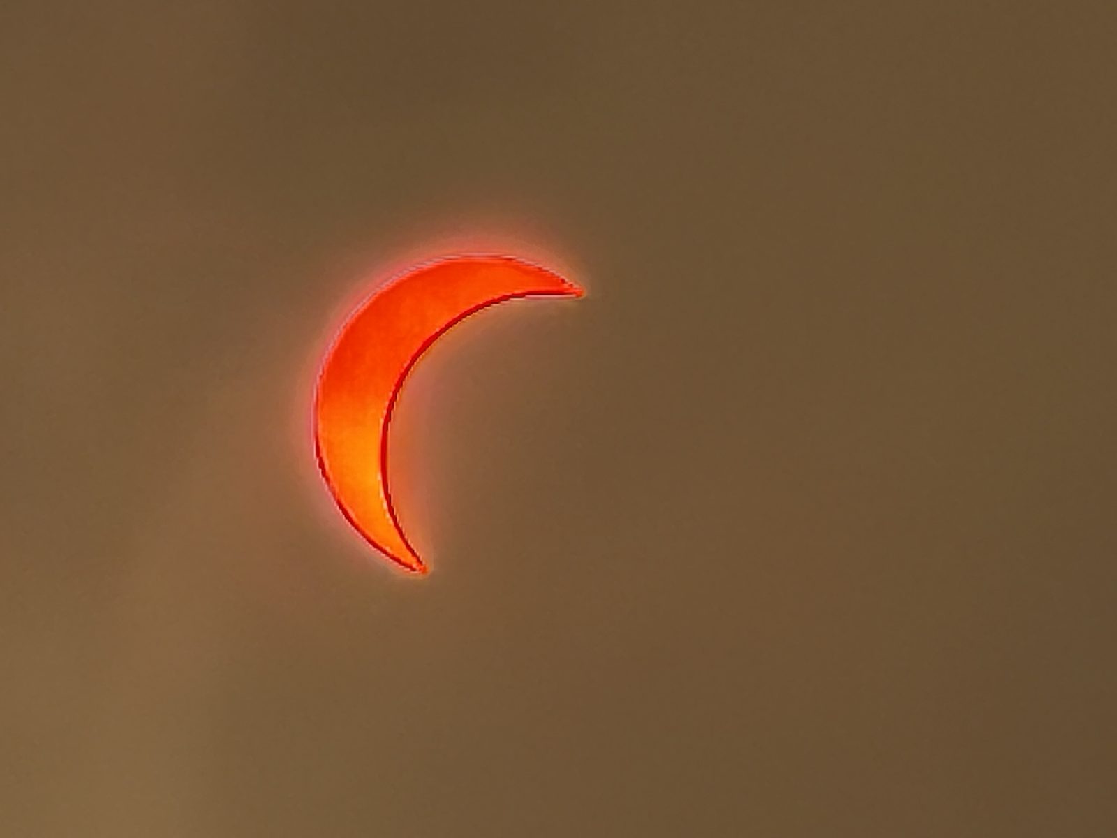 A partial solar eclipse seen as a deep red crescent through a sky thick with wildfire smoke