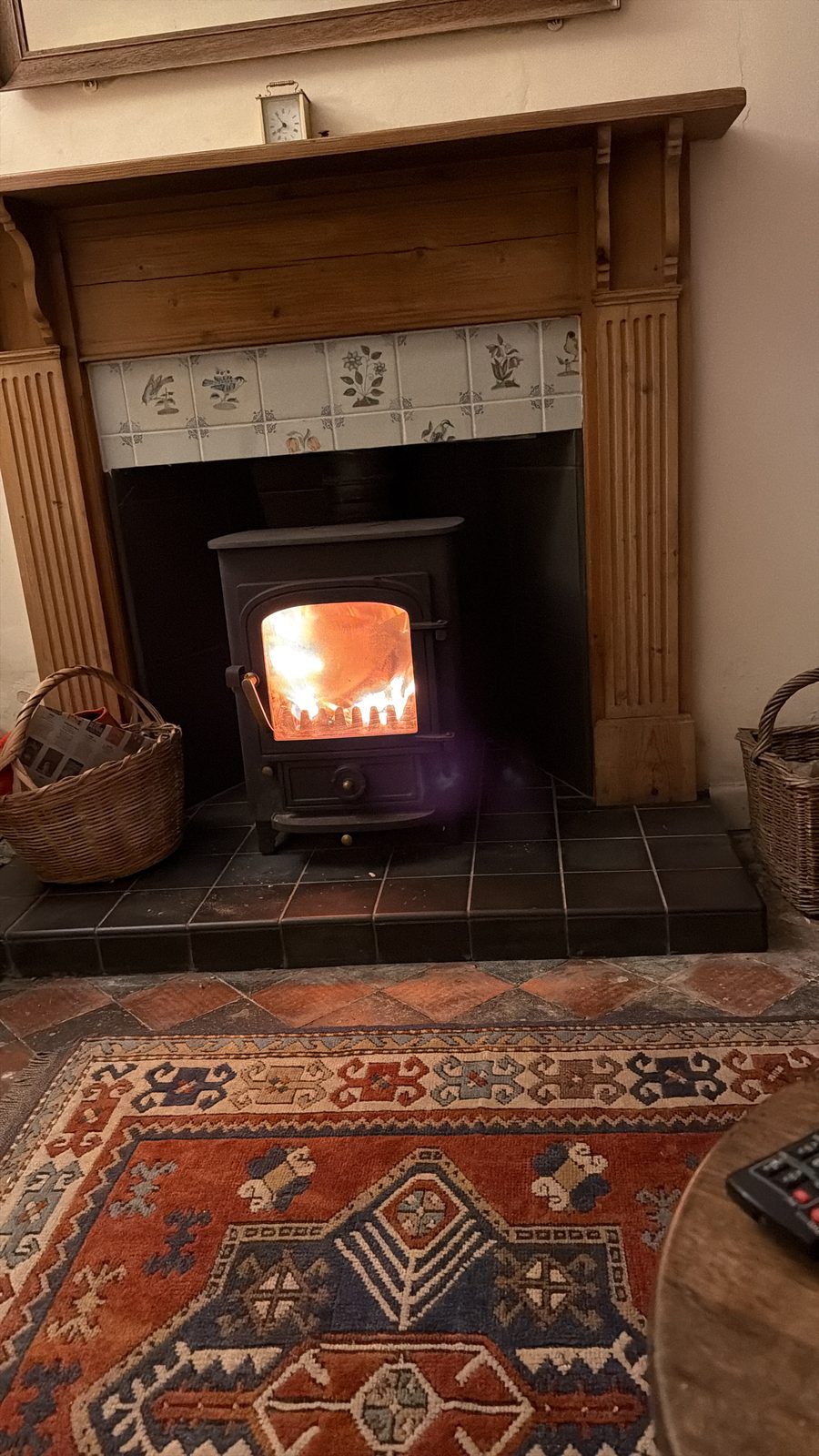 A wood-burning stove alight in the pine fireplace surround at No. 2, with decorative tiles, a kilim rug and log baskets