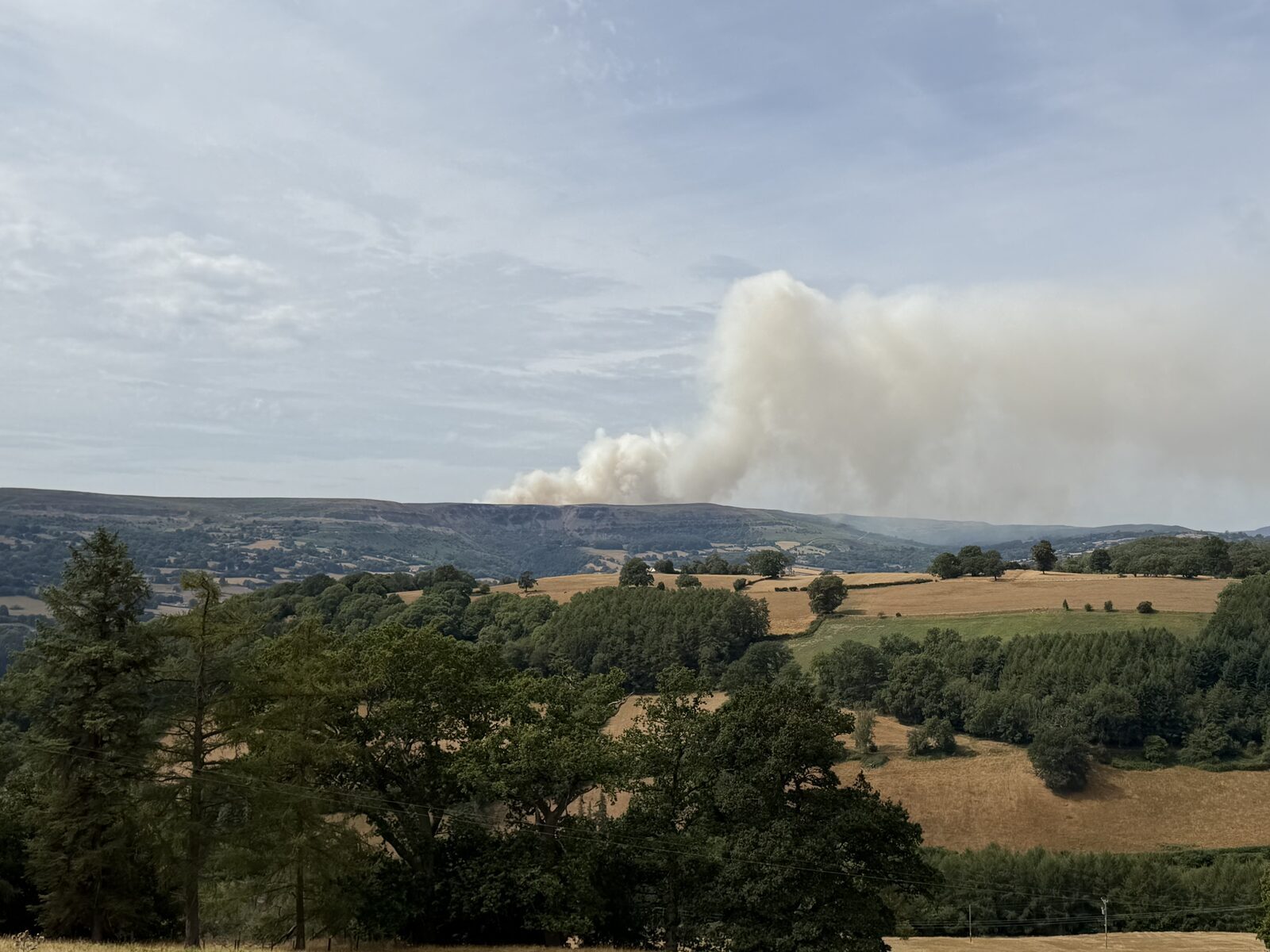 A large plume of pale smoke rising from a hillside fire on the far side of the valley, above Llangattock, seen across parched summer fields and wooded slopes from Pengilfach