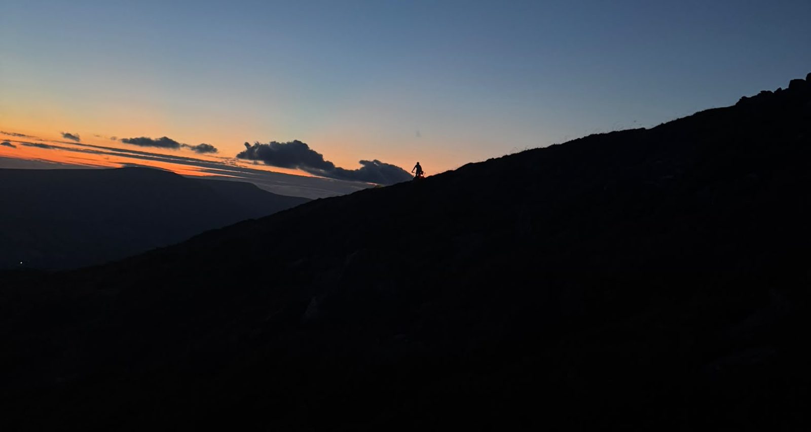 A lone mountain biker silhouetted on the ridge of the Sugarloaf against a deep orange sunset, riding home down the mountain
