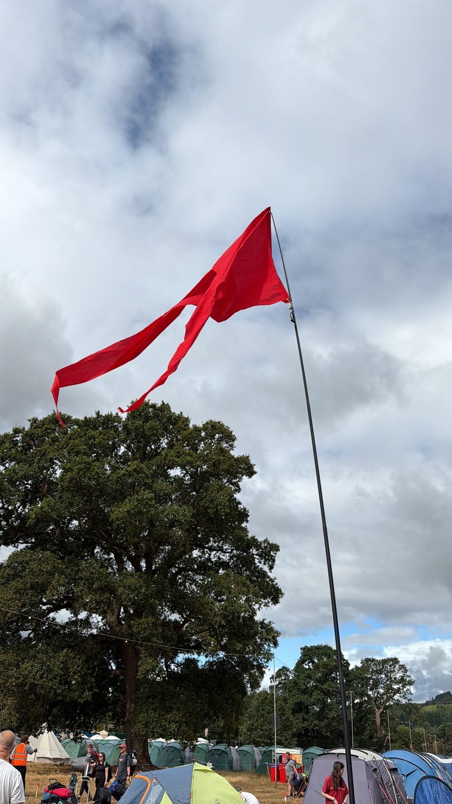 A tall red pennant flag — the family camp flag — flying from a pole high above the festival campsite, with a big oak tree and rows of tents below a cloudy sky