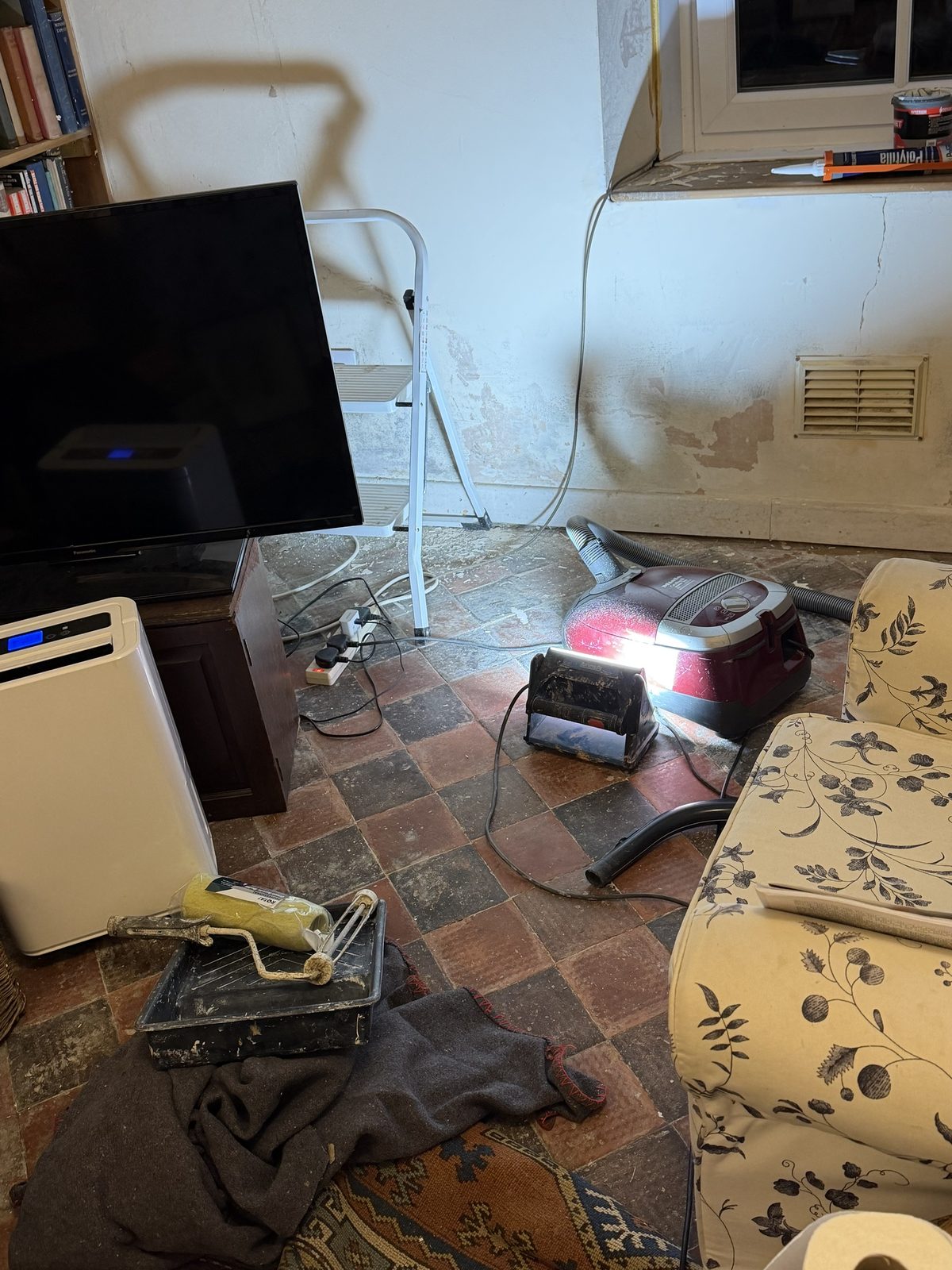 The sitting room mid-decoration, a step ladder, paint tray and roller, vacuum and dust sheets on the quarry-tiled floor