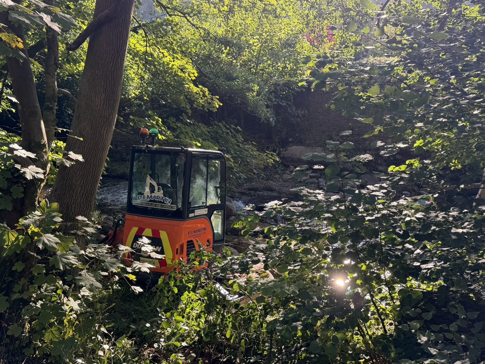 An orange mini-digger working down in the wooded riverbed, dappled sunlight through the trees