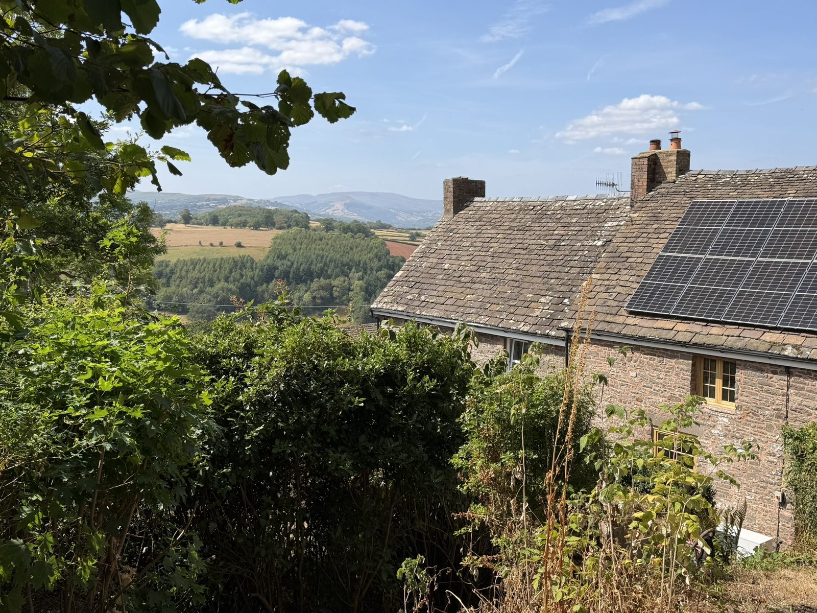 A summer view over the valley and distant hills from the rear of the cottages, the stone cottage with its slate roof and solar panels to one side, framed by greenery