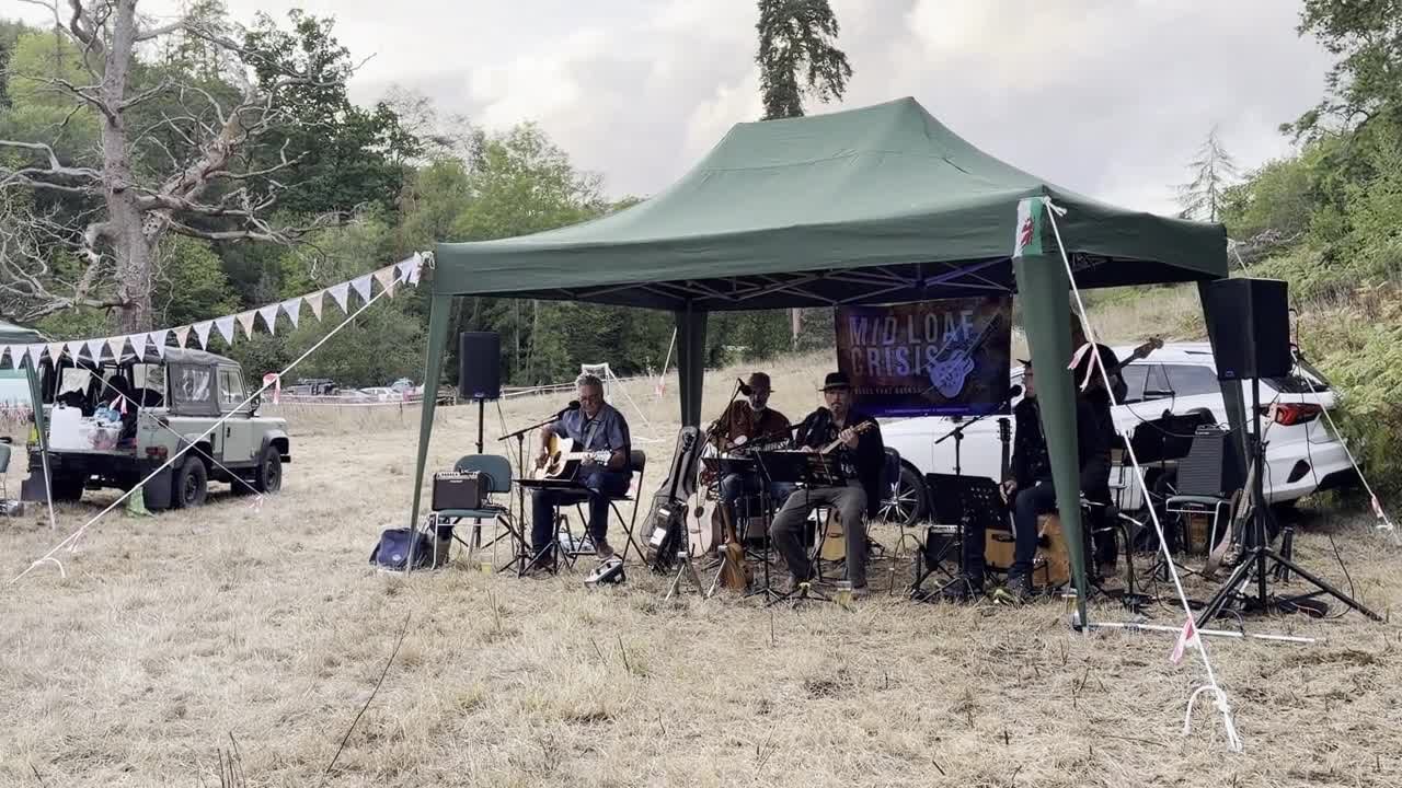 A band playing under a green gazebo at the village party, guitars and instruments, bunting strung across the field