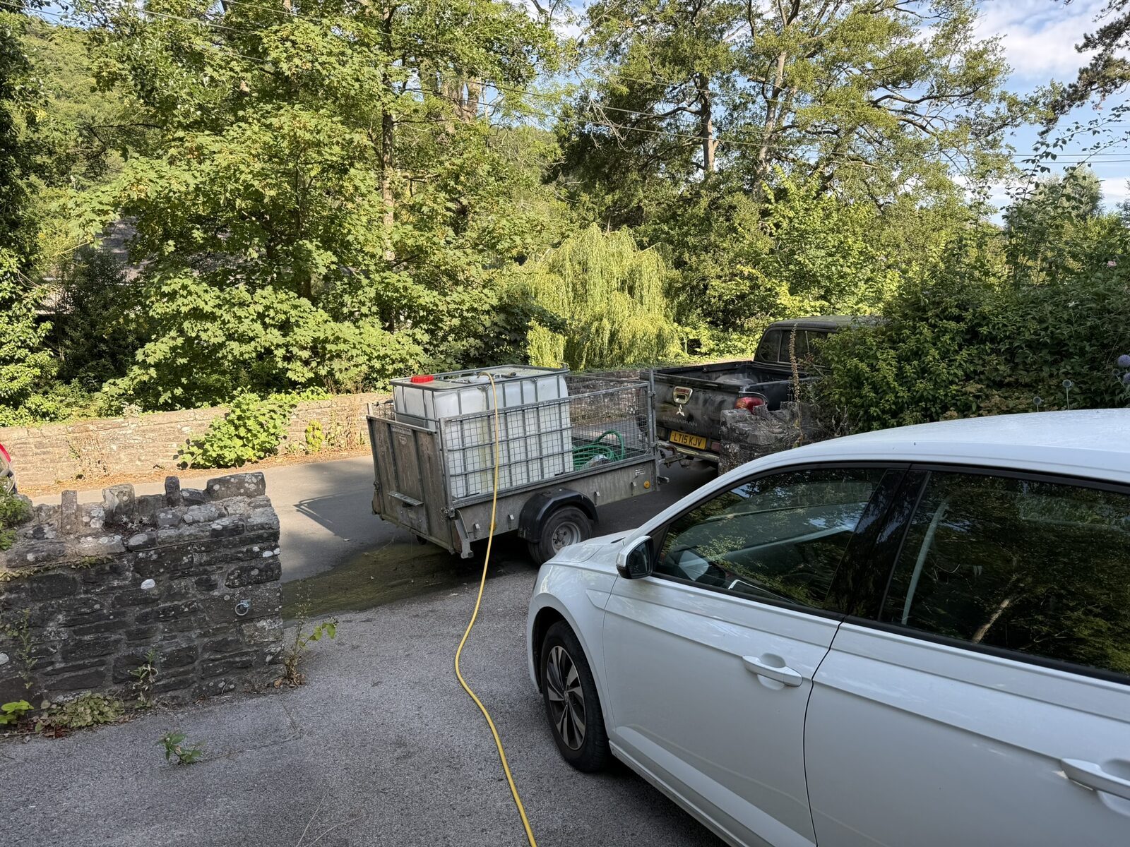A 1,000-litre white IBC water tank on a trailer down by the road, a yellow hosepipe running to it from a kind neighbour's mains supply, stone wall and trees behind
