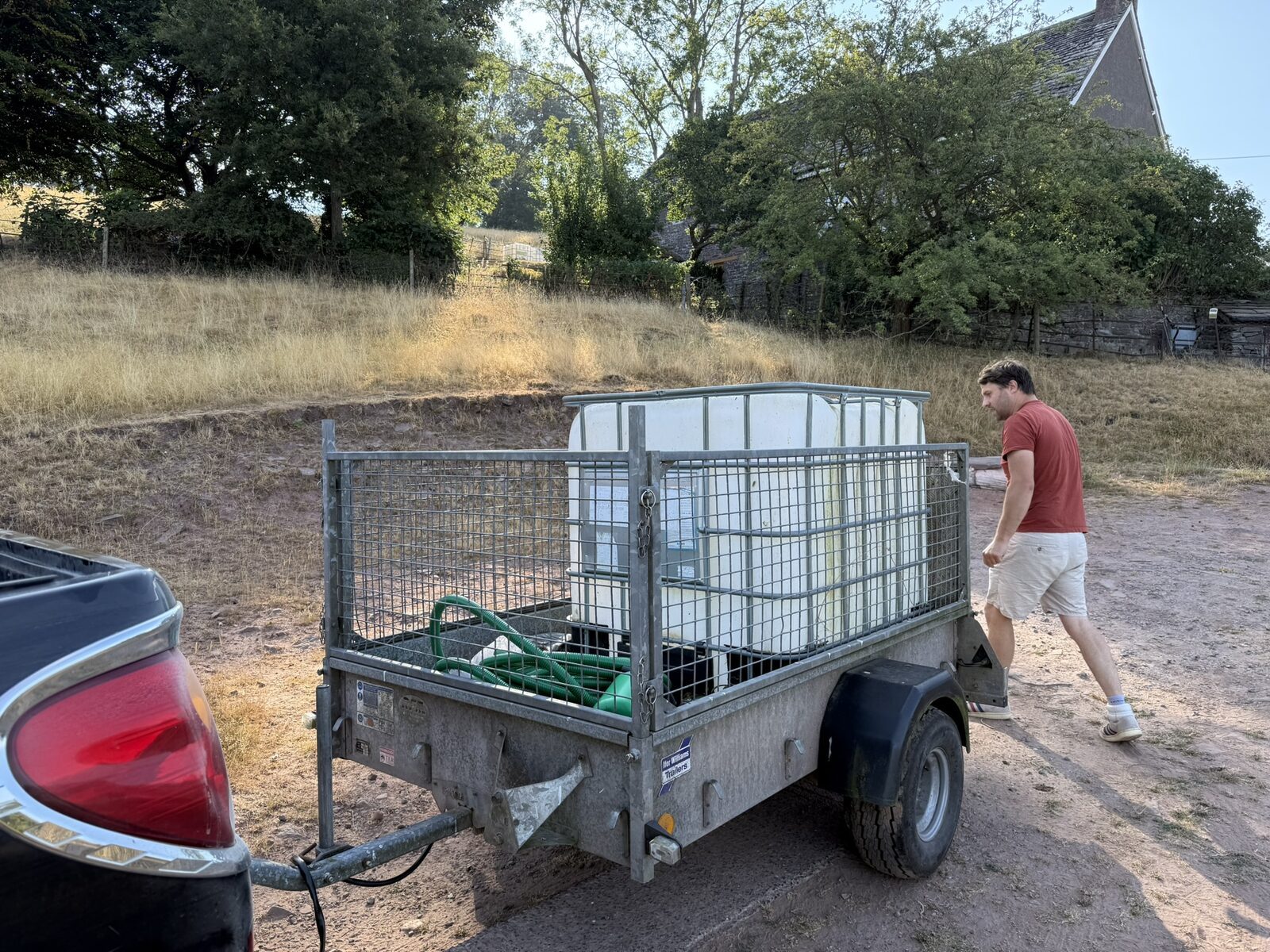 A 1,000-litre white IBC water tank in a steel cage, loaded in a mesh-sided trailer on the gravel parking area at the cottages, with Tom Jayne walking past