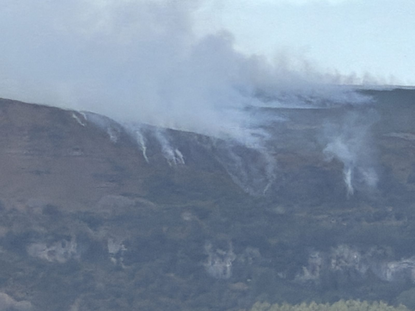 Close view of the wildfire burning along the top of Llangattock mountain, pale smoke rising from several points on the blackened hillside
