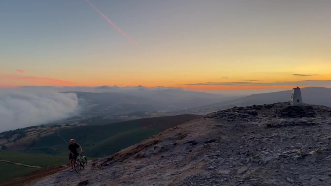 From the Sugarloaf summit at sunset, a wall of smoke rolling down into the valley as a cyclist climbs the ridge