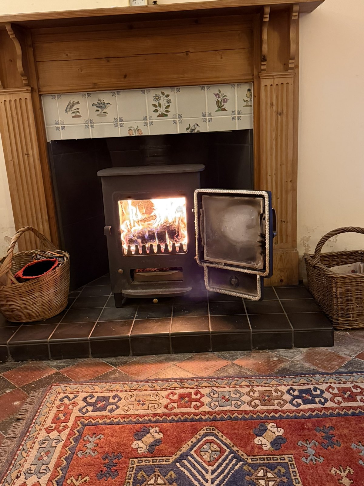 A wood-burning stove alight in a pine fireplace with hand-painted bird and flower tiles above, log baskets either side and a kilim rug on the quarry-tiled floor